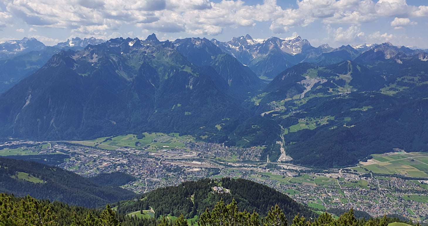 Ein Panoramablick auf eine Talstadt, umgeben von grünen Hügeln und fernen schneebedeckten Bergen unter einem teilweise bewölkten Himmel.