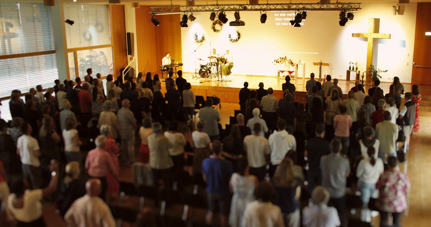 Eine Gemeinde steht in einer Kirche und blickt zu den Musikern auf der Bühne, die neben einem großen Kreuz und einer Leinwand mit Liedtexten stehen.