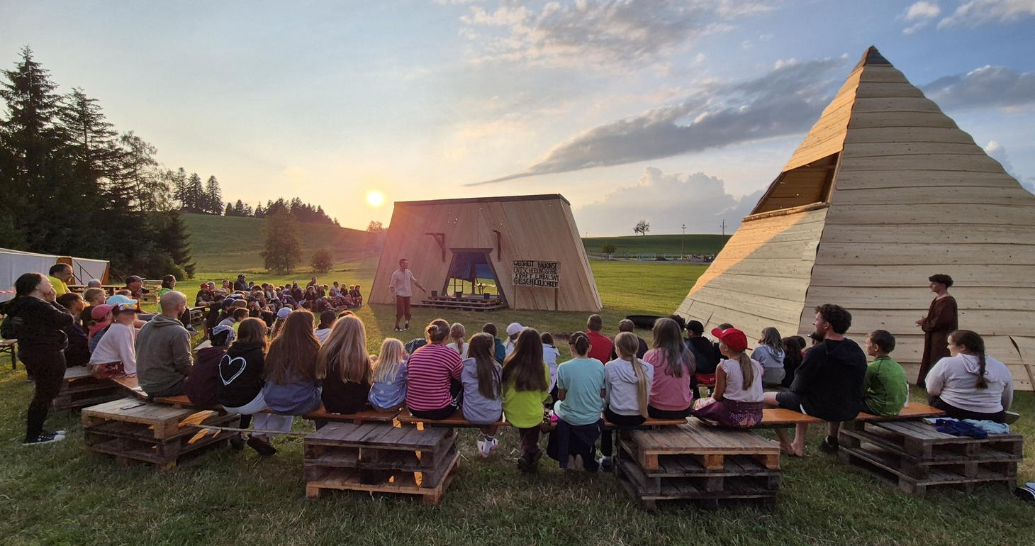 Kinder sitzen auf Bänken und beobachten bei Sonnenuntergang einen Künstler im Freien in der Nähe moderner Holzkonstruktionen.