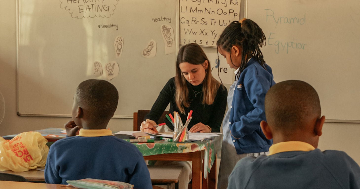 Eine Lehrerin sitzt an einem Tisch und hilft einem Schüler, während zwei andere Kinder in der Nähe in einem Klassenzimmer sitzen.
