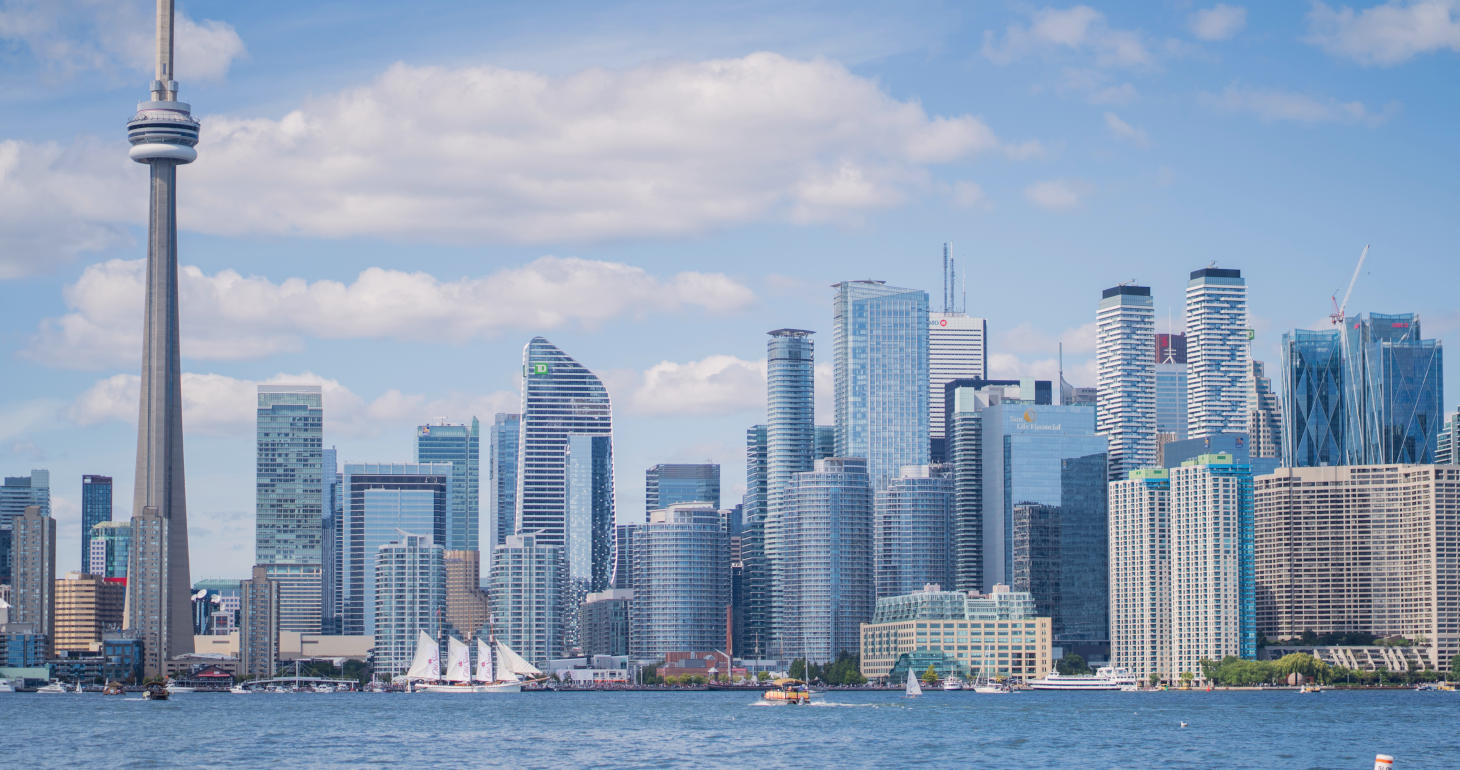 Die Skyline von Toronto mit hohen Gebäuden, dem CN Tower, Segelbooten und blauem Himmel mit Wolken, vom anderen Ufer aus gesehen.