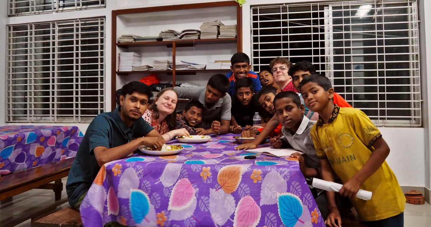 Eine Gruppe von Jugendlichen und ein Erwachsener sitzen um einen Tisch mit Essen herum und lächeln in einem hellen Raum in die Kamera.