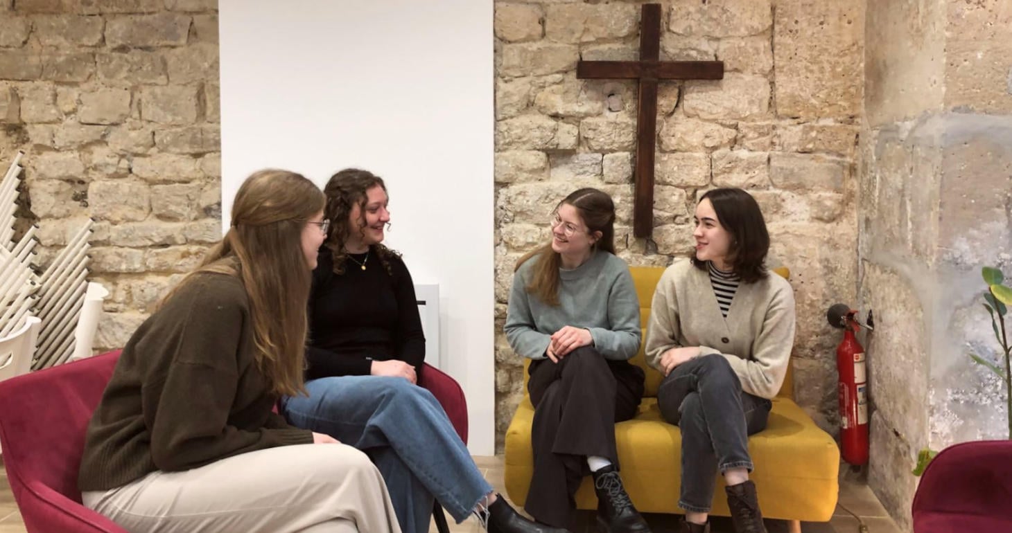 Vier Frauen sitzen auf Stühlen und einer gelben Couch in einem Raum mit Steinwänden und einem Holzkreuz an der Wand und unterhalten sich.