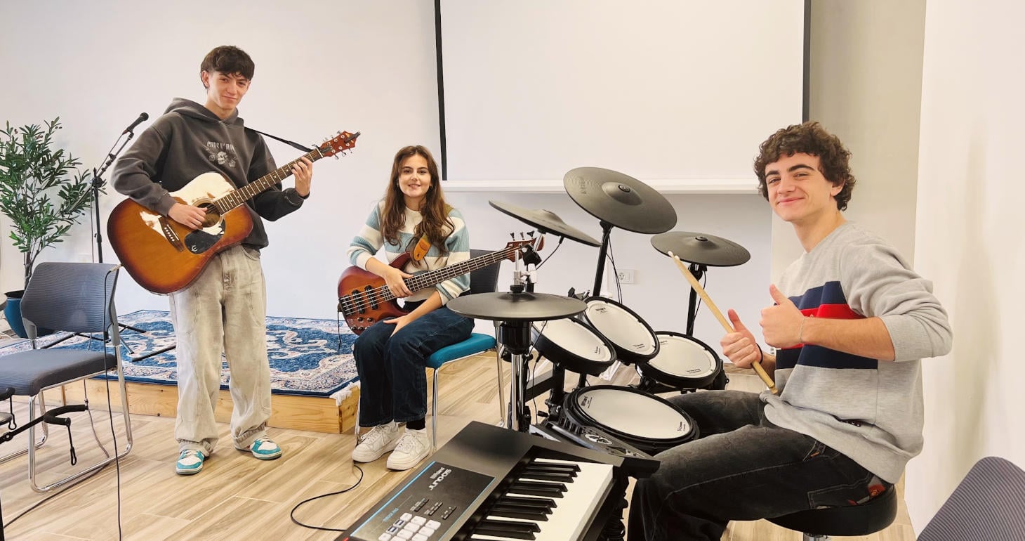 Drei lächelnde Teenager spielen in einem hellen Raum Gitarre, Bass und Schlagzeug, während einer neben einem Keyboard den Daumen hochhält.