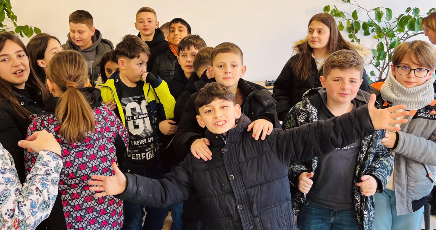 Eine Gruppe von Kindern steht lächelnd zusammen in einem Raum, wobei ein Junge ganz vorne steht und die Arme weit ausbreitet.