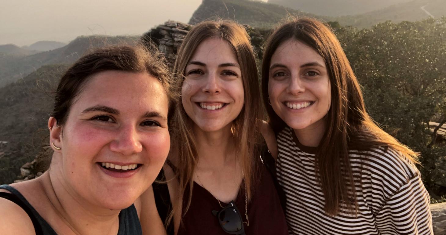 Drei junge Frauen, die im Freien lächeln, im Hintergrund sind bei Sonnenuntergang Berge und Bäume zu sehen.