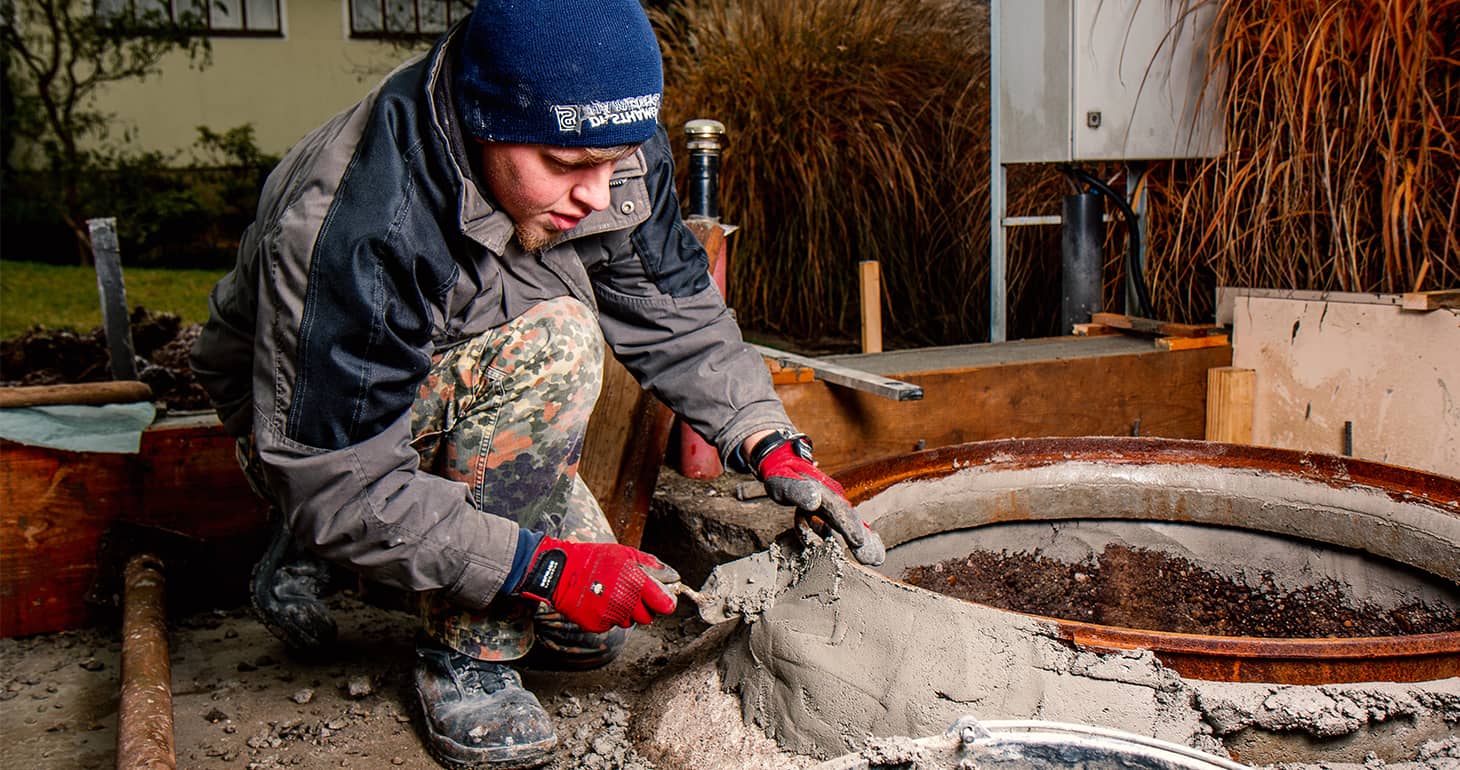 Ein Arbeiter mit Handschuhen und einer Mütze glättet im Freien nassen Beton um eine runde Metallschalung herum.