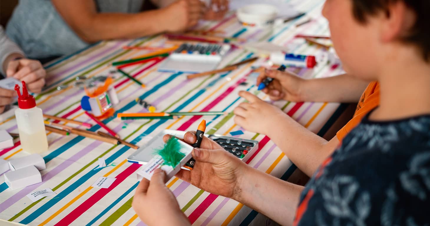 Kinder, die auf einer gestreiften Tischdecke mit Buntstiften, Papier und anderem Bastelmaterial basteln.
