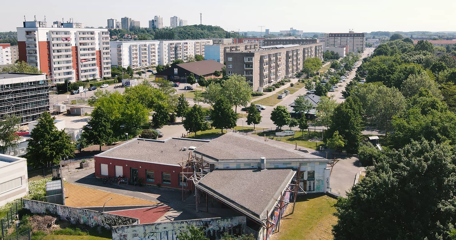 Luftaufnahme eines Wohngebiets mit Mehrfamilienhäusern, grünen Bäumen und einigen niedrigen Gebäuden im Vordergrund.