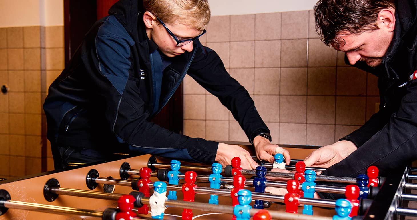Zwei Personen spielen konzentriert Tischfußball und sind ganz auf ihr Spiel vertieft; sie befinden sich in einem Innenraum mit hellbraunen Fliesenwänden.