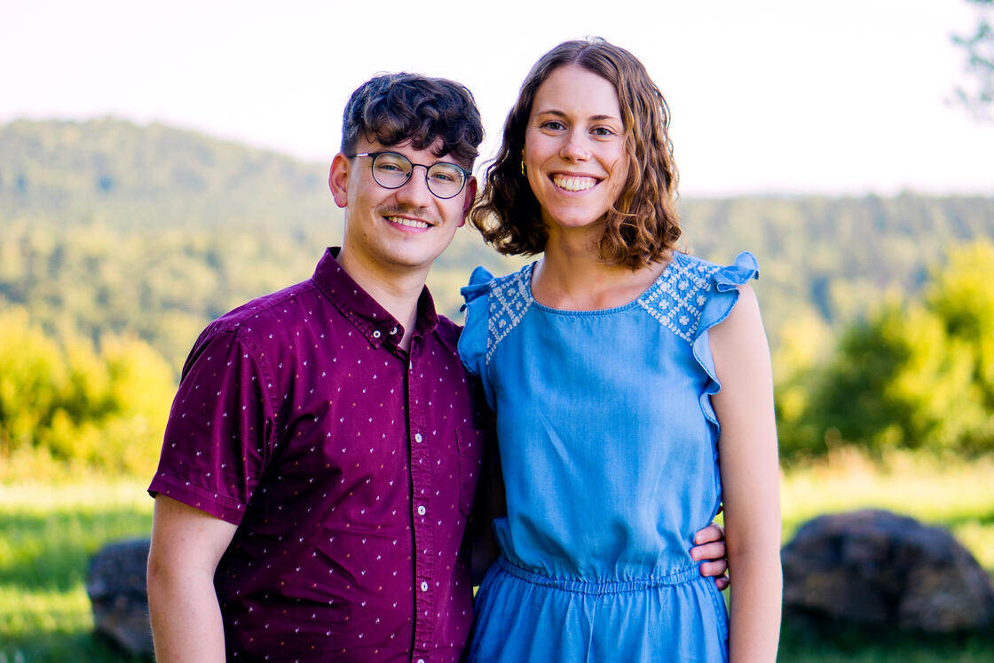 Ein lächelnder Mann in einem weinroten Hemd und eine Frau in einem blauen Kleid stehen gemeinsam im Freien, hinter ihnen sind grüne Bäume zu sehen.