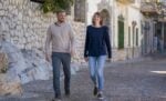 Ein Mann und eine Frau gehen gemeinsam auf einer Kopfsteinpflasterstraße neben einer Steinmauer entlang, lächeln und unterhalten sich.
