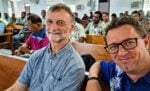 Zwei lächelnde Männer sitzen in der ersten Reihe eines überfüllten Raums mit Holzbänken und Menschen im Hintergrund.