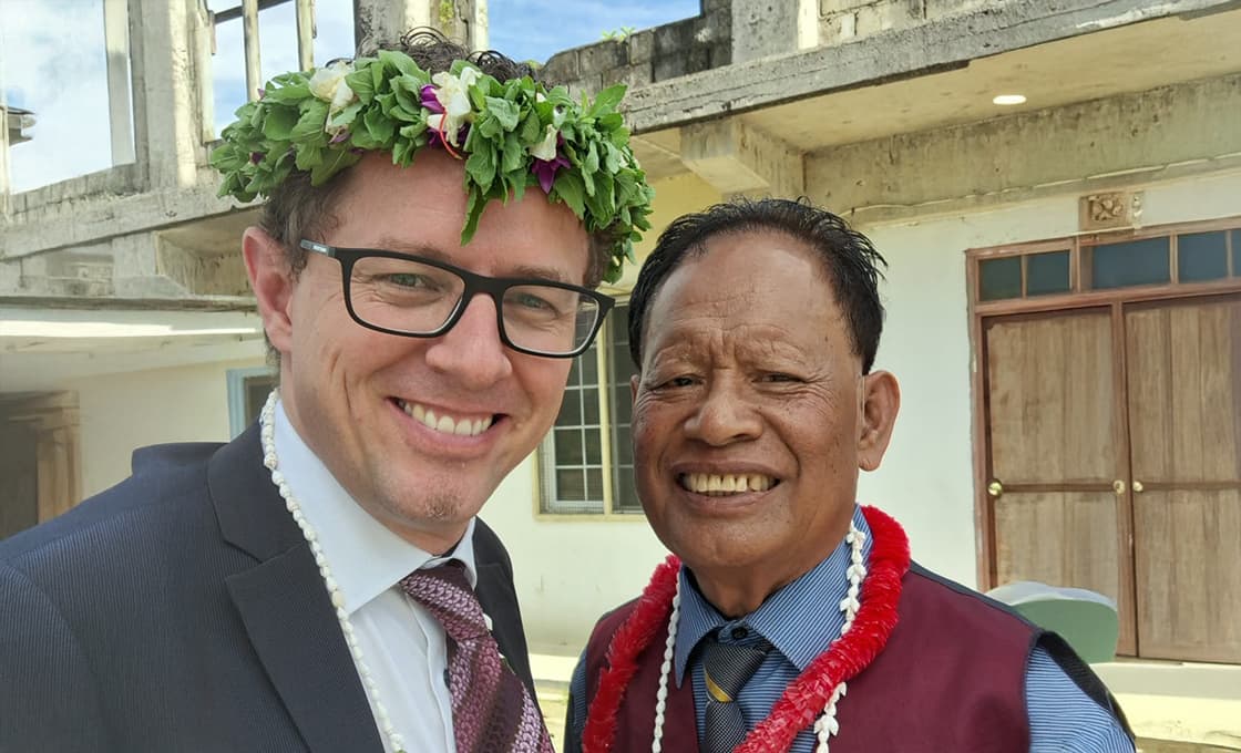 Zwei Männer lächeln im Freien, beide tragen Anzüge und Blumenketten; einer trägt eine Brille und einen Blumenkranz. Hinter ihnen steht ein Betongebäude.