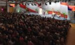 Ein Redner spricht zu einem sitzenden Publikum in einem großen Saal mit einer Bühne, Stühlen und einem „LETS GO!“-Schild.