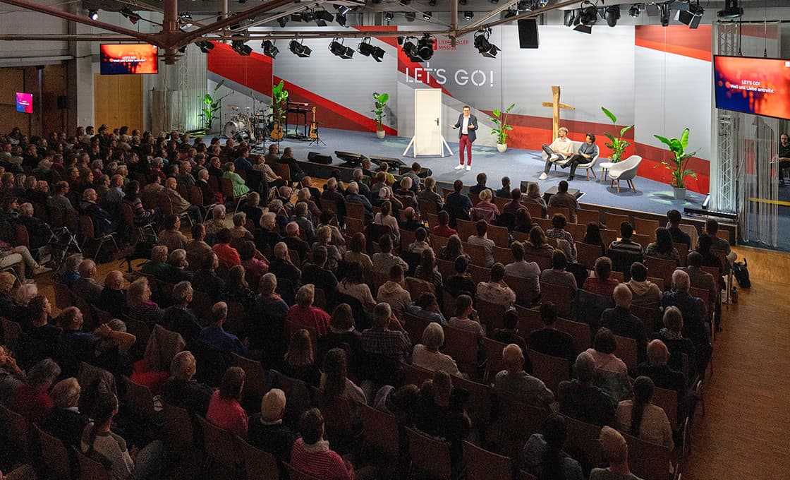 Ein Redner spricht zu einem sitzenden Publikum in einem großen Saal mit einer Bühne, Stühlen und einem „LETS GO!“-Schild.