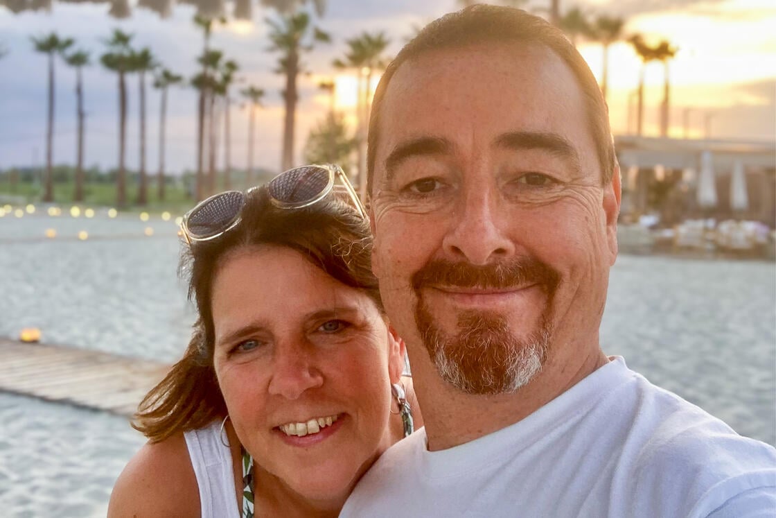 Ein lächelndes Paar, das bei Sonnenuntergang am Strand ein Selfie macht, im Hintergrund sind Palmen und ein Gebäude zu sehen.