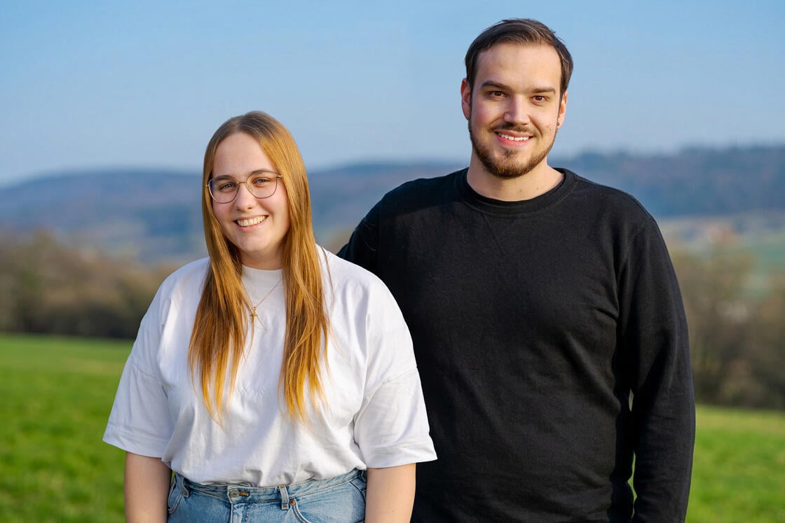 Zwei Menschen stehen lächelnd auf einer Wiese, im Hintergrund sind Hügel und ein blauer Himmel zu sehen.