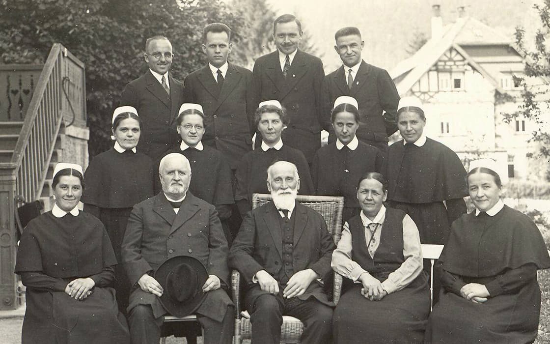 Eine Gruppe von Krankenschwestern und Männern in festlicher Kleidung posiert vor einem großen Haus für ein Schwarz-Weiß-Foto.
