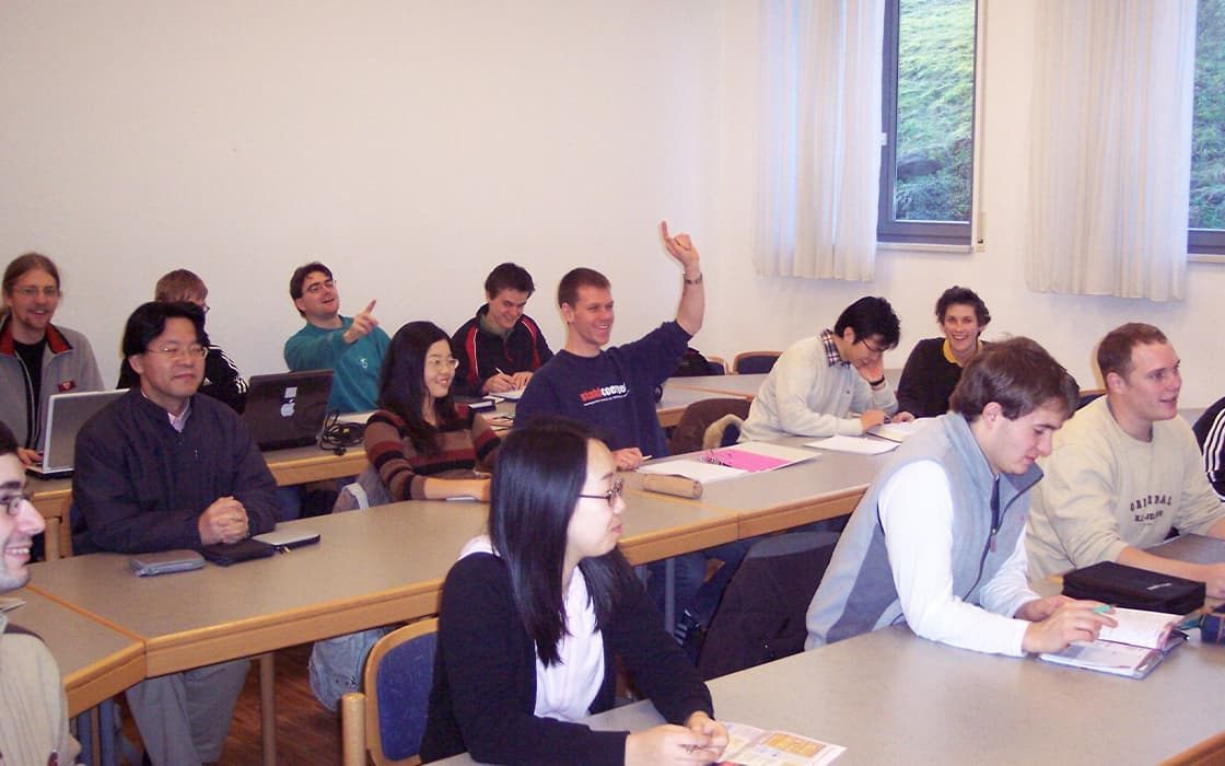 Schüler, die an Schreibtischen in einem Klassenzimmer sitzen, einige lächeln, einer hebt die Hand, im Hintergrund sind Fenster zu sehen.