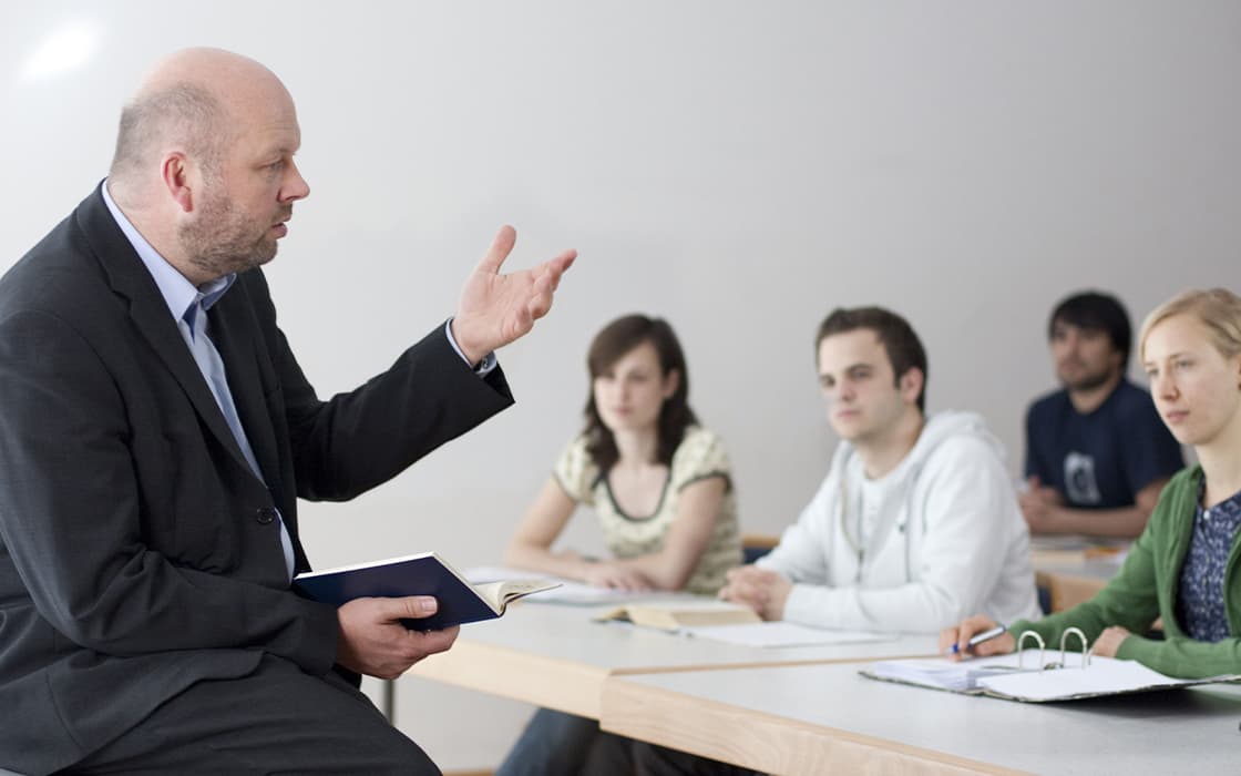 Ein Lehrer im Anzug gestikuliert, während er mit vier Schülern spricht, die an ihren Schreibtischen in einem Klassenzimmer sitzen.