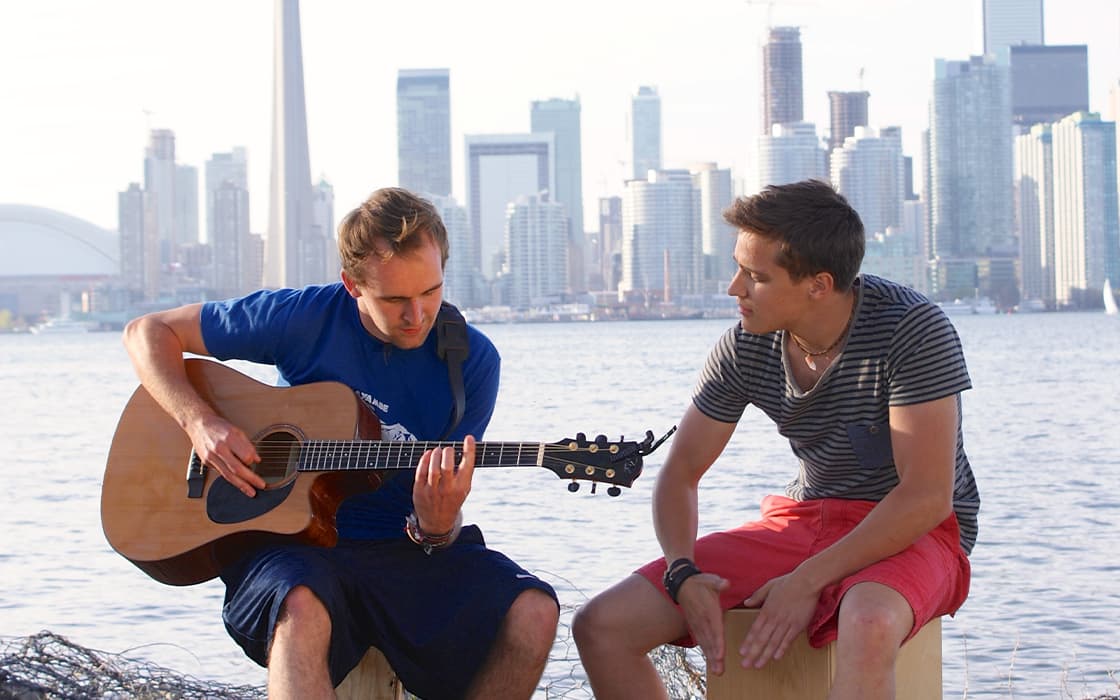 Zwei junge Männer sitzen am Wasser, einer spielt Gitarre, im Hintergrund sind die Skyline der Stadt und hohe Gebäude zu sehen.