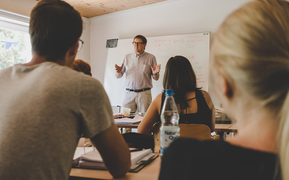 Ein Lehrer steht vorne im Klassenzimmer und spricht zu den Schülern, die mit ihren Heften an den Schreibtischen sitzen.