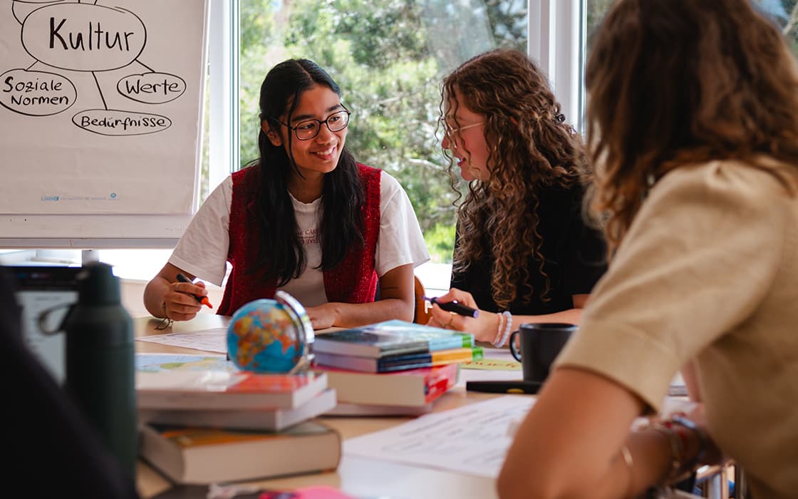 Drei Schüler sitzen mit Büchern an einem Tisch, unterhalten sich und lächeln; daneben stehen eine Whiteboard-Tafel und ein Globus.