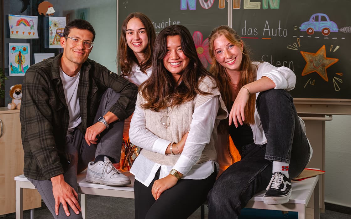 Vier junge Erwachsene, die gemeinsam in einem Klassenzimmer lächeln und posieren, im Hintergrund eine bunte Tafel.