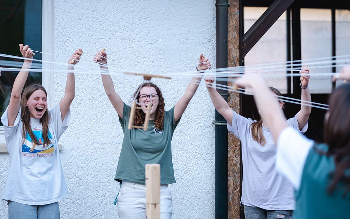 Drei Personen im Freien heben die Arme und halten dabei Schnüre fest – eine Teambildungsübung mit einer Holzkonstruktion.