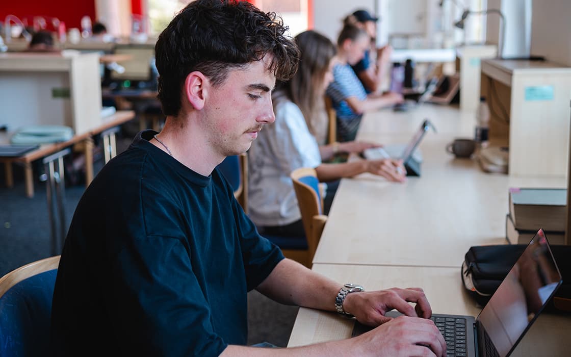 Eine Person tippt an einem langen Schreibtisch in einer Bibliothek auf einem Laptop, während im Hintergrund andere arbeiten.