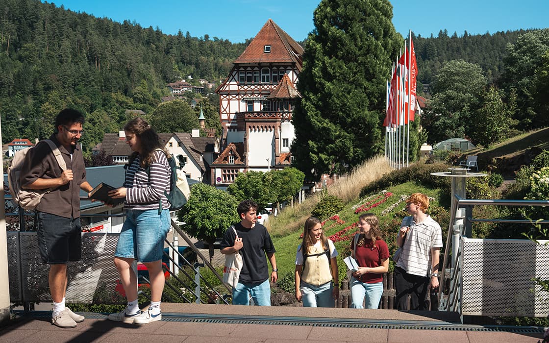 Eine Gruppe von Schülern geht draußen spazieren und unterhält sich, im Hintergrund sind Bäume, Fahnen und traditionelle Gebäude zu sehen.