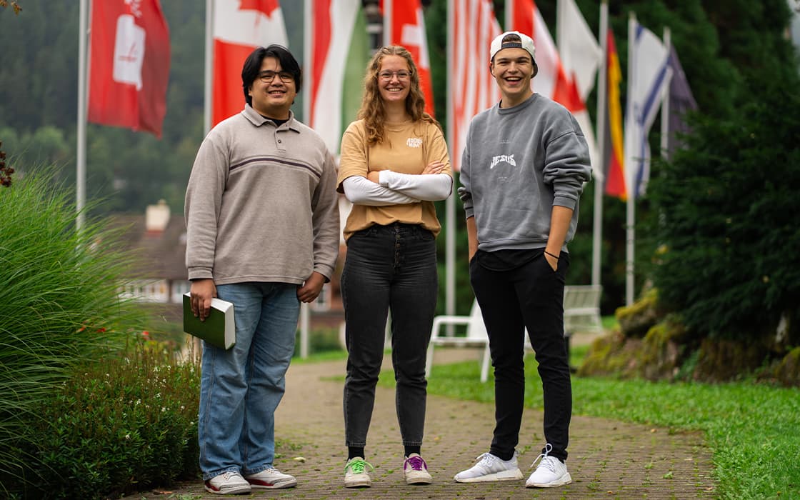 An einem bewölkten Tag stehen drei Menschen lächelnd auf einem Weg, im Hintergrund sind internationale Flaggen zu sehen.