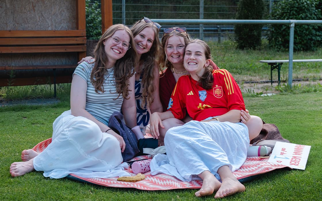 Vier lächelnde Frauen sitzen eng beieinander auf einer Decke im Freien und genießen an einem sonnigen Tag ein Picknick.