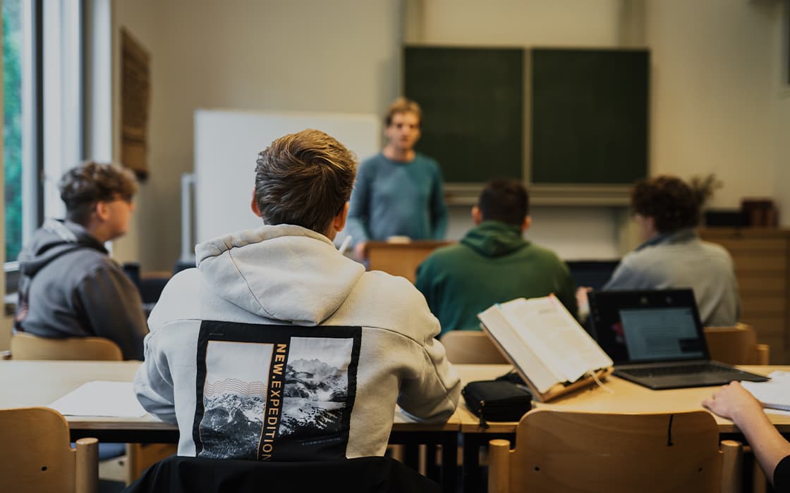 Die Schüler sitzen an ihren Schreibtischen und blicken auf einen Lehrer in einem Klassenzimmer mit einer grünen Tafel und aufgeschlagenen Büchern.