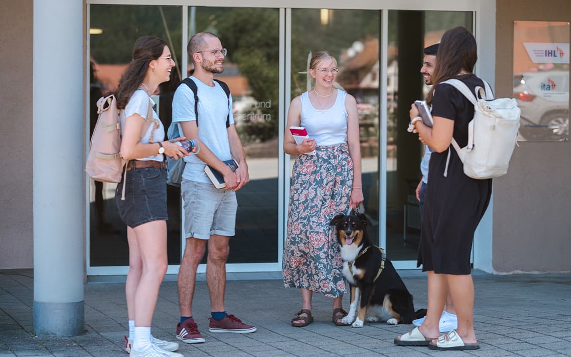Fünf Menschen und ein Hund stehen vor einem Gebäude, lächeln und unterhalten sich an einem sonnigen Tag.
