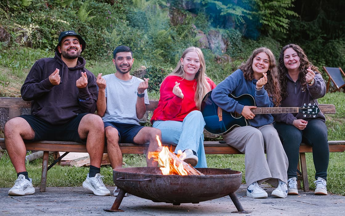 Fünf Freunde sitzen lächelnd am Lagerfeuer, einer spielt Gitarre, und alle formen mit den Fingern ein Herz.