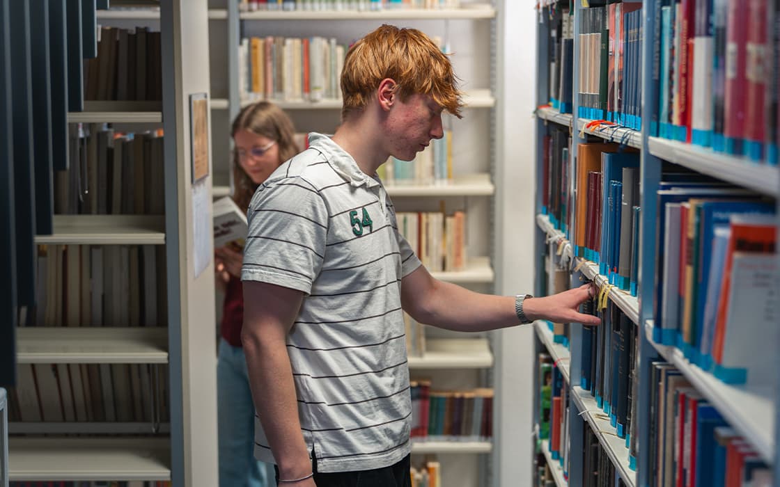 Ein junger Mann in einem gestreiften Hemd stöbert in den Bücherregalen einer Bibliothek, während im Hintergrund eine Frau liest.