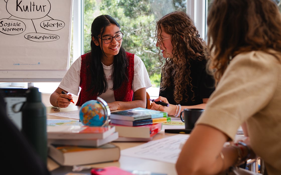 Drei Schüler sitzen um einen Tisch herum und lernen gemeinsam; im Hintergrund ist eine Tafel zu sehen, auf der deutsche Begriffe stehen.