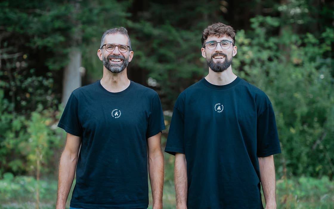 Zwei Männer in schwarzen T-Shirts stehen lächelnd nebeneinander im Freien, im Hintergrund ist Grün zu sehen.