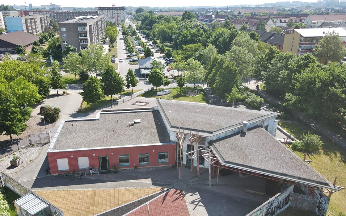 Luftaufnahme eines modernen Gebäudes, umgeben von Bäumen, mit einer Stadtstraße und Wohnhäusern im Hintergrund.