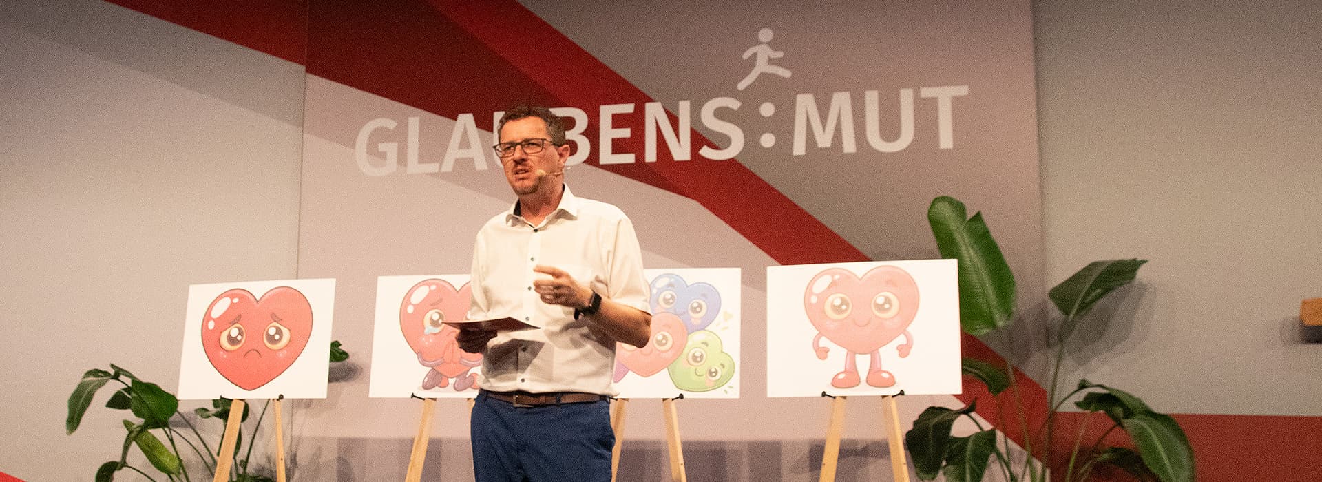 Ein Mann spricht auf einer Bühne, im Hintergrund sind Plakate mit Herzmotiven im Cartoon-Stil und Pflanzen zu sehen.