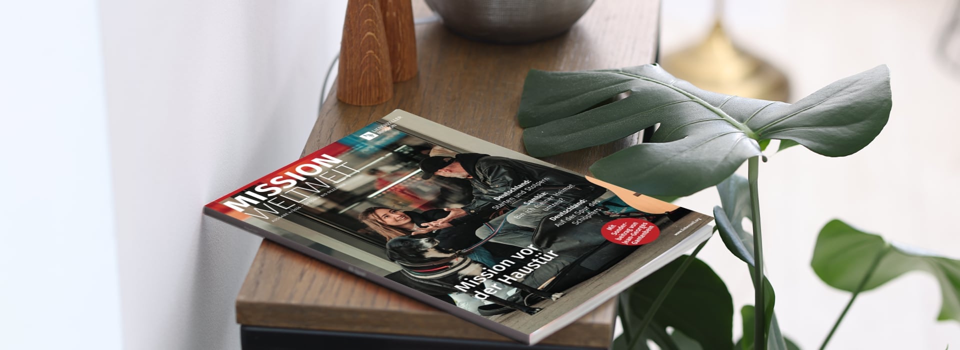 Die Zeitschrift „Mission Weltweit“ liegt auf einem Holztisch neben einer grünen Pflanze und Dekoartikeln.