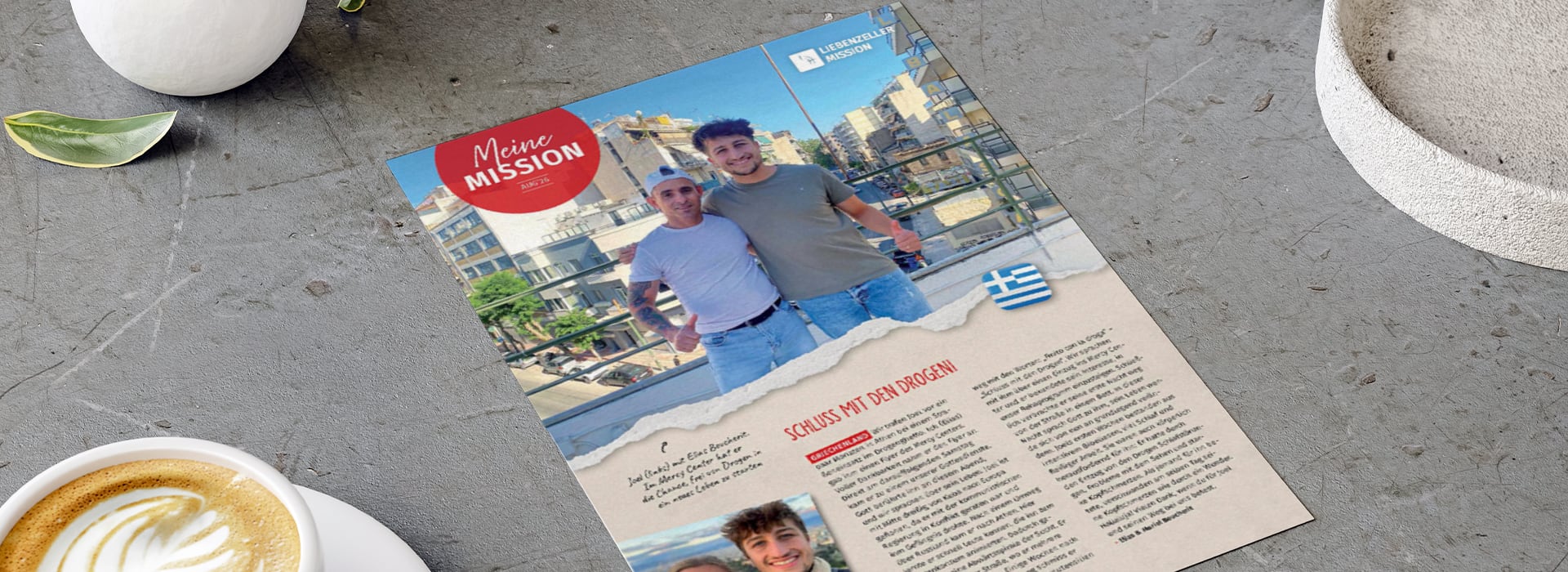 Auf einer Zeitschriftenseite, die auf einem Tisch liegt, sind zwei lächelnde Männer im Freien zu sehen; neben ihrem Foto befinden sich ein Text und eine griechische Flagge.