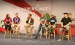 Auf der Bühne sitzen fünf Personen auf Hockern und unterhalten sich, während im Hintergrund ein „Glaubens:Mut“-Banner zu sehen ist.