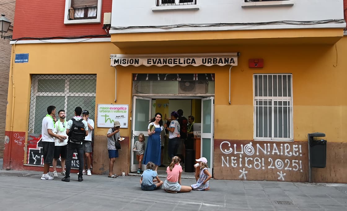 Vor einem gelben Gebäude mit der Aufschrift „Misión Evangélica Urbana“ stehen und sitzen Menschen. Kinder sitzen auf dem Bürgersteig.