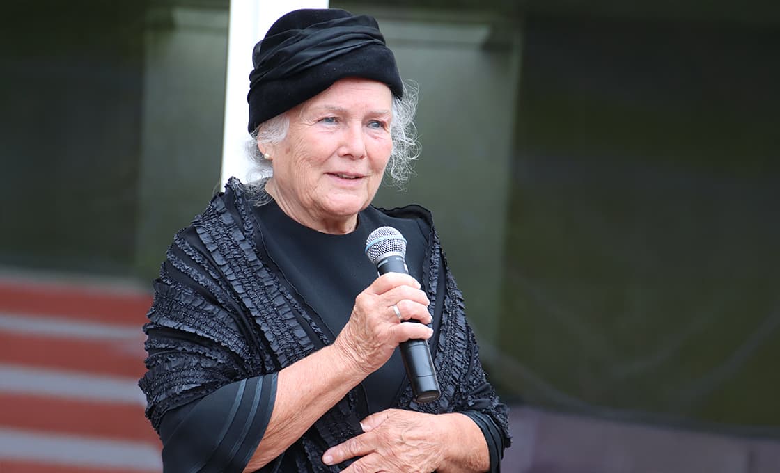 Judi Dench trägt einen schwarzen Hut und einen Schal und hält ein Mikrofon in der Hand, während sie spricht.