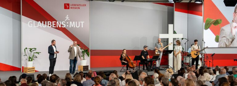 Zwei Männer stehen auf einer Bühne vor einer Band und einem Publikum, hinter ihnen ist an der Wand der Schriftzug GLAUBENS:MUT und ein Kreuz zu sehen.