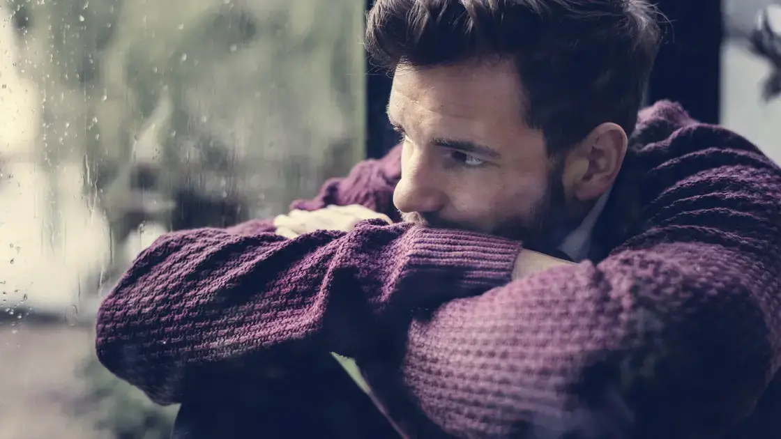 Ein Mann in einem lila Pullover, der sein Kinn auf seine verschränkten Arme stützt, während er aus einem regennassen Fenster schaut.