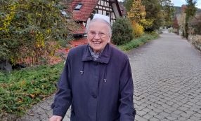 Schwester Gertrud lächelt in einem dunklen Mantel auf einem Kopfsteinpflasterweg und feiert ihren 100. Geburtstag inmitten von Häusern und Grünanlagen.